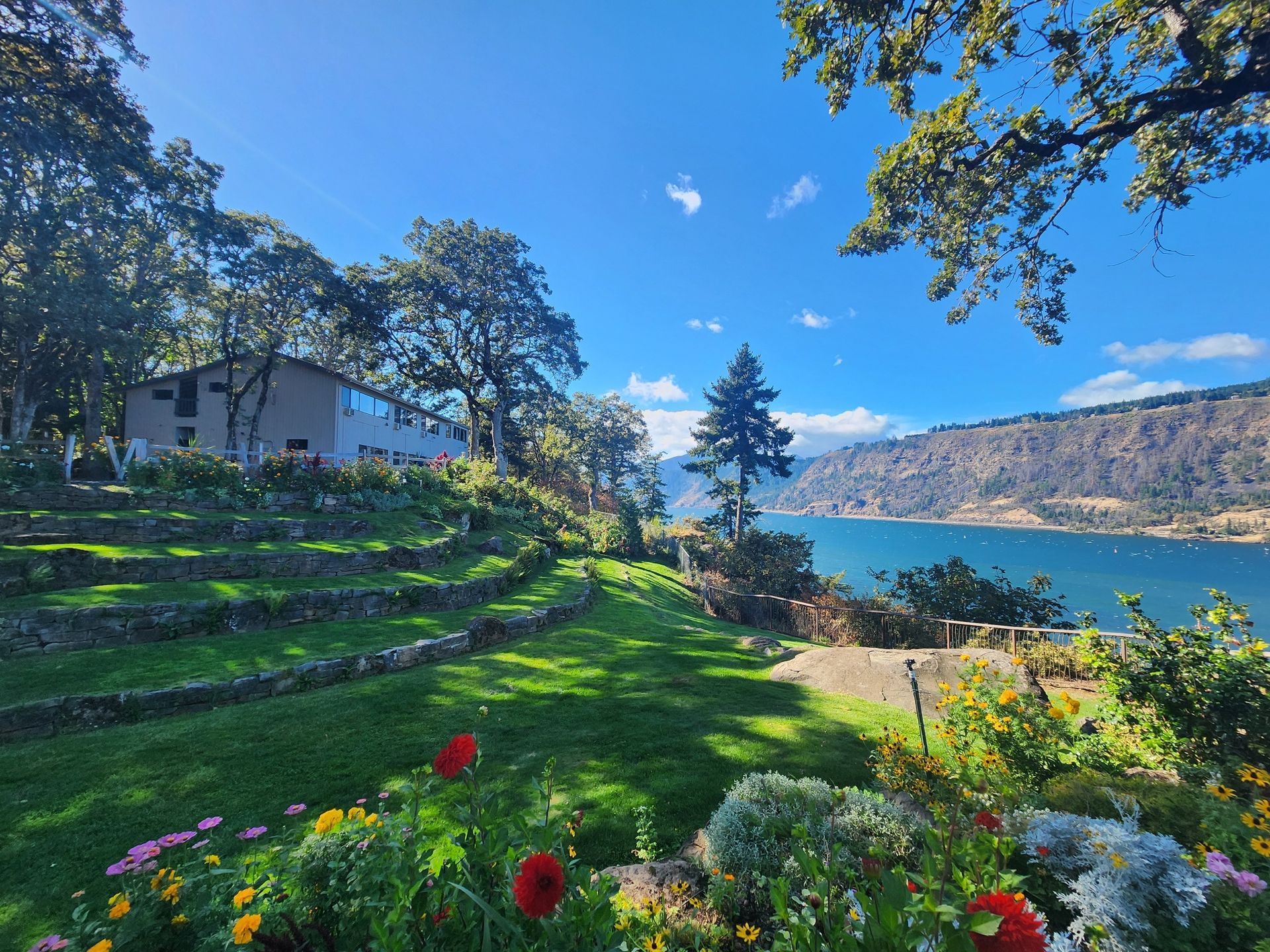 Terraced garden with flowers, trees, and a building overlooking a lake and mountains under a blue sky.