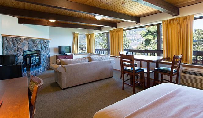 Hotel room with a stone fireplace, beige sofa, wooden table, and a bed. Brown beams and yellow curtains.