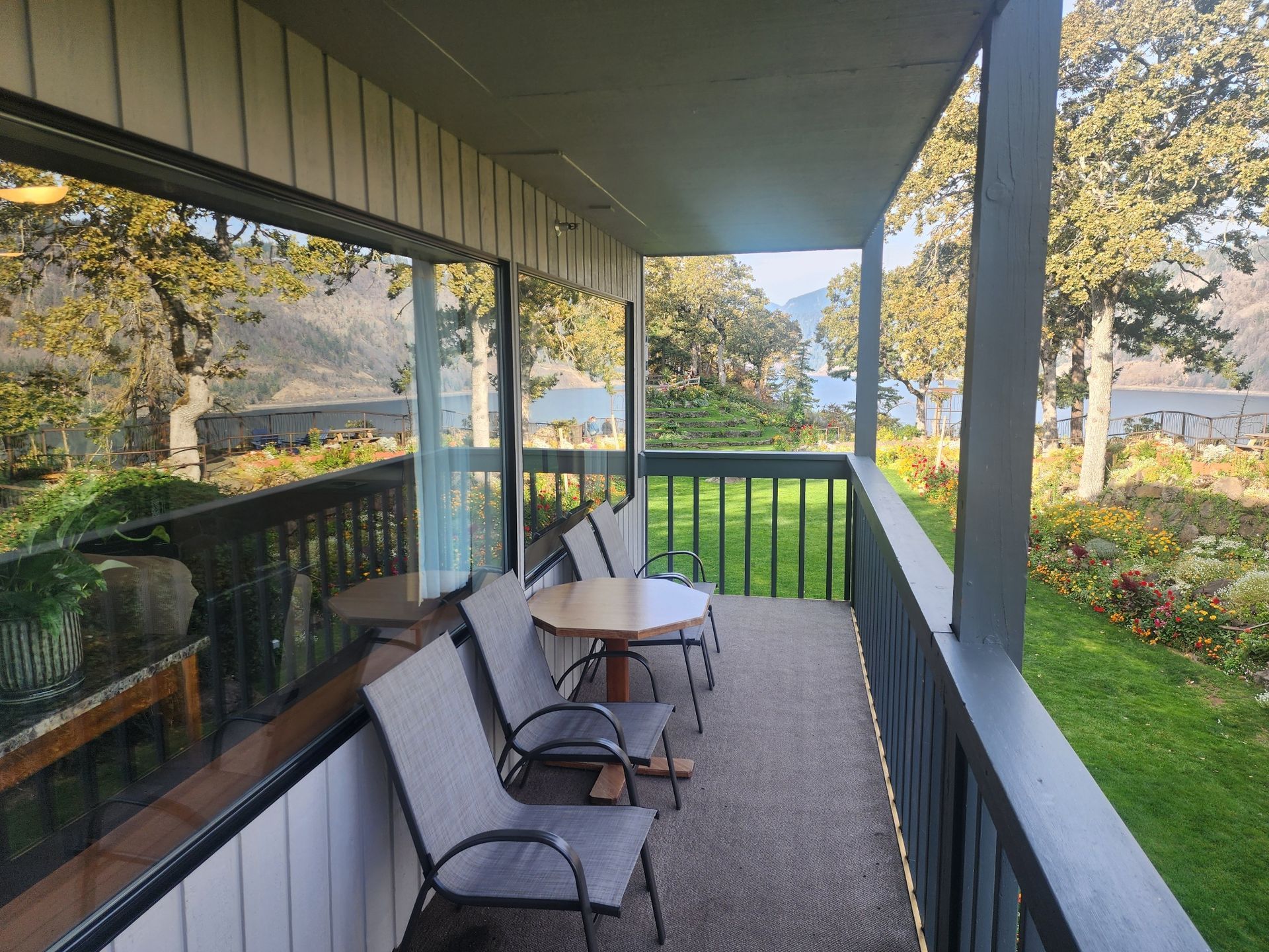 Balcony with table, chairs, glass wall, and view of trees, water, and building.