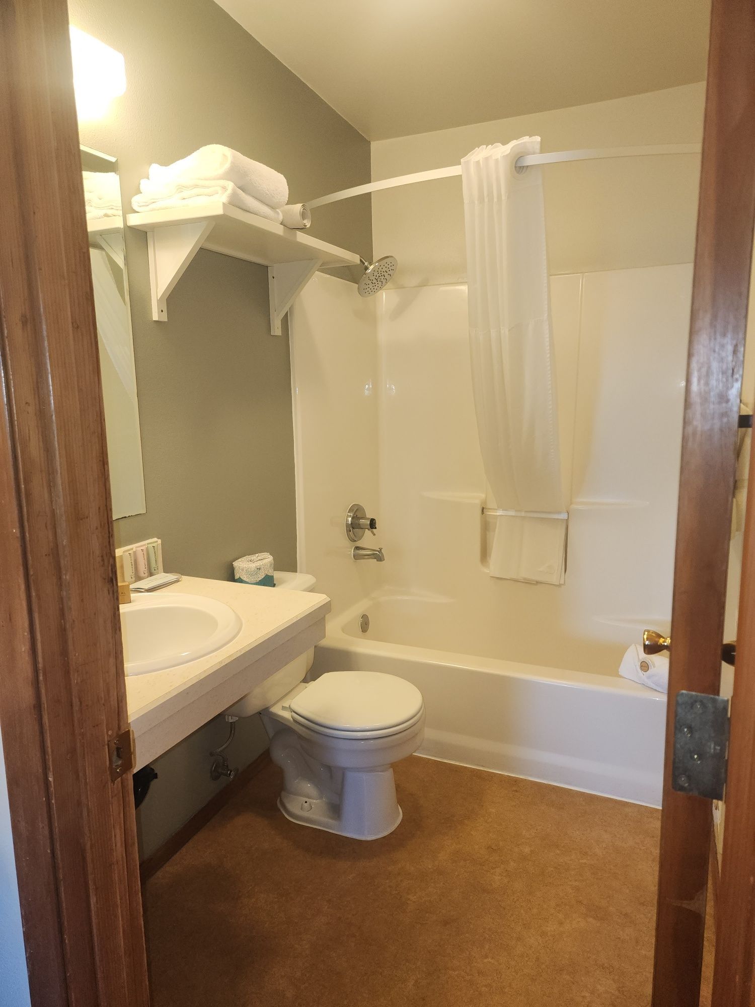 Bathroom with a white bathtub, toilet, and sink. Towels are stacked on shelves. Brown carpet and beige walls.