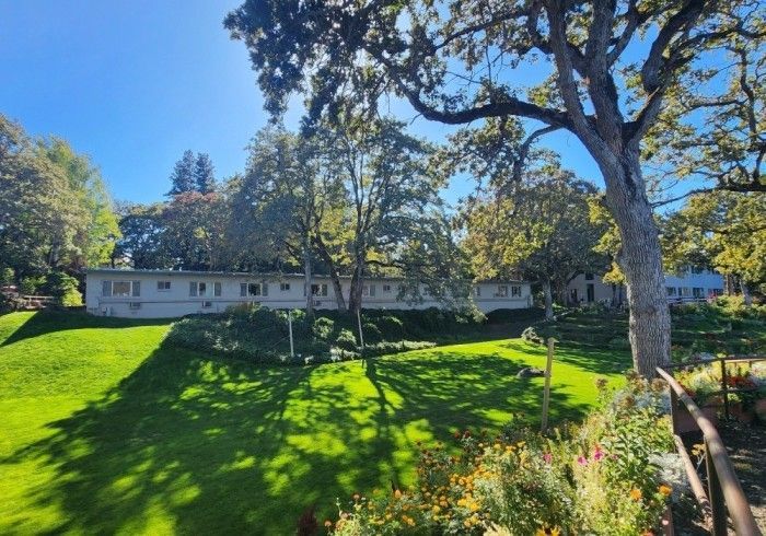 A long, single-story building sits behind a green lawn and trees under a blue sky.