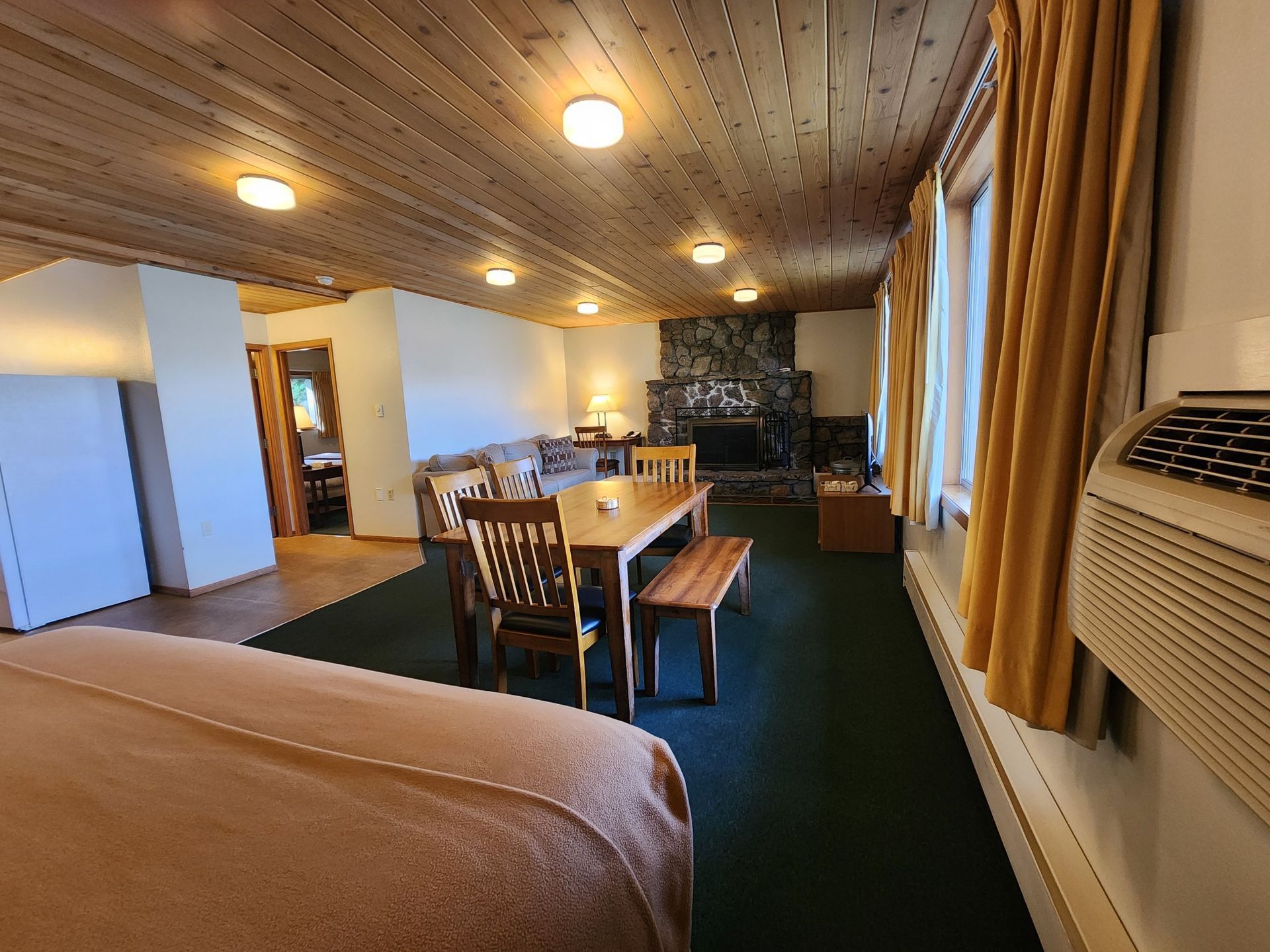 Spacious room with dining table, fireplace, and bed. Wooden ceiling and yellow curtains.