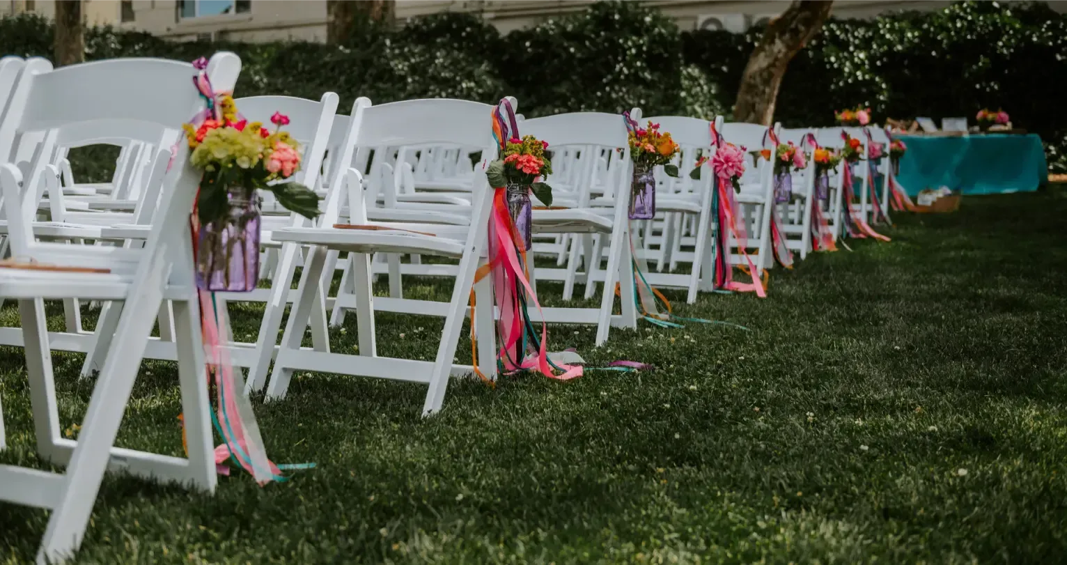 White folding chairs decorated with colorful ribbons and flowers, arranged on grass.
