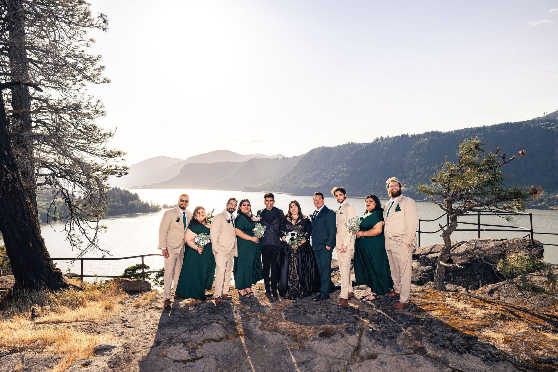 Wedding party on a cliff overlooking a lake. People are in emerald and tan attire with the couple in black.