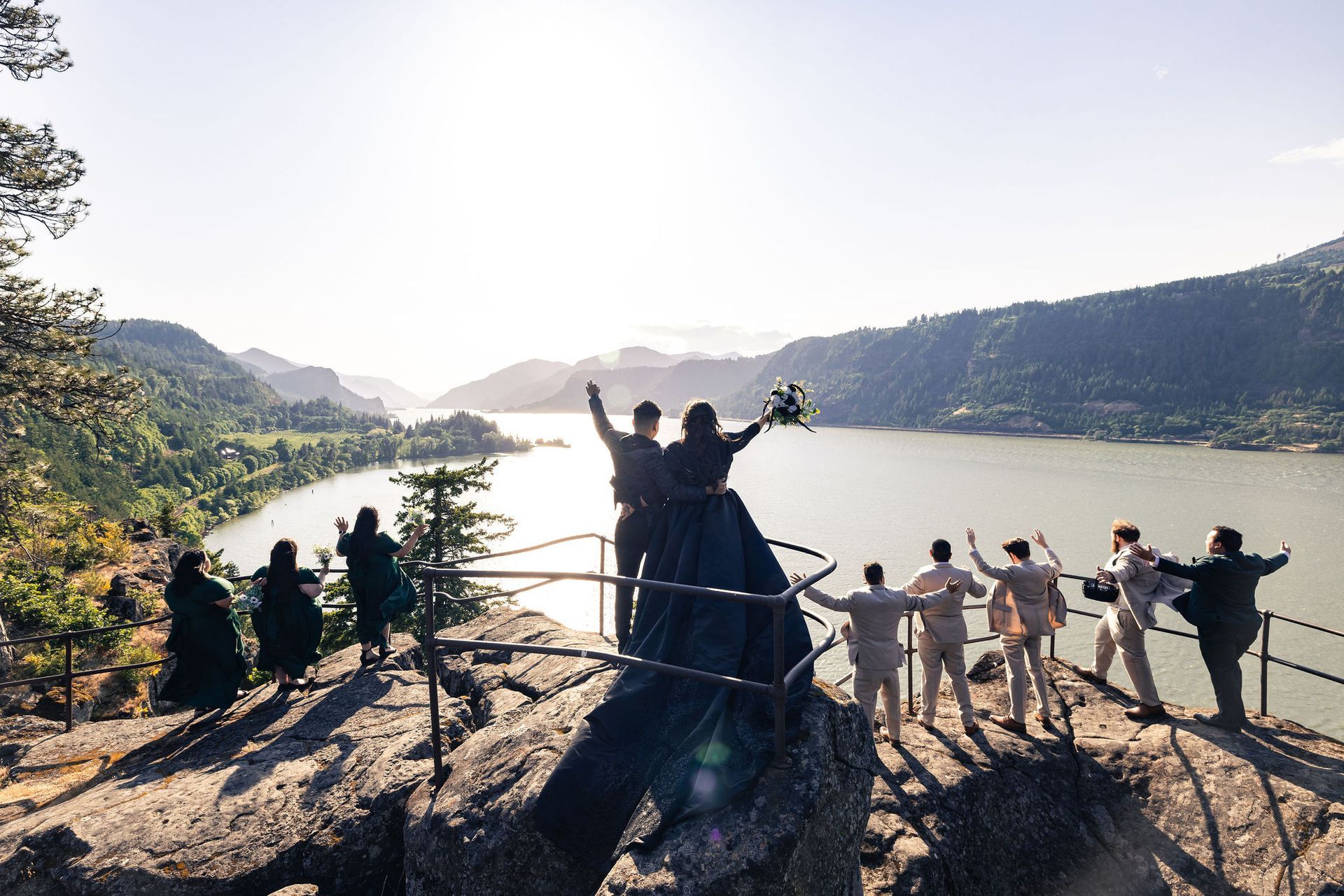 Wedding party on a rocky cliff overlooking a lake and mountains, arms raised in celebration. Sunny day.