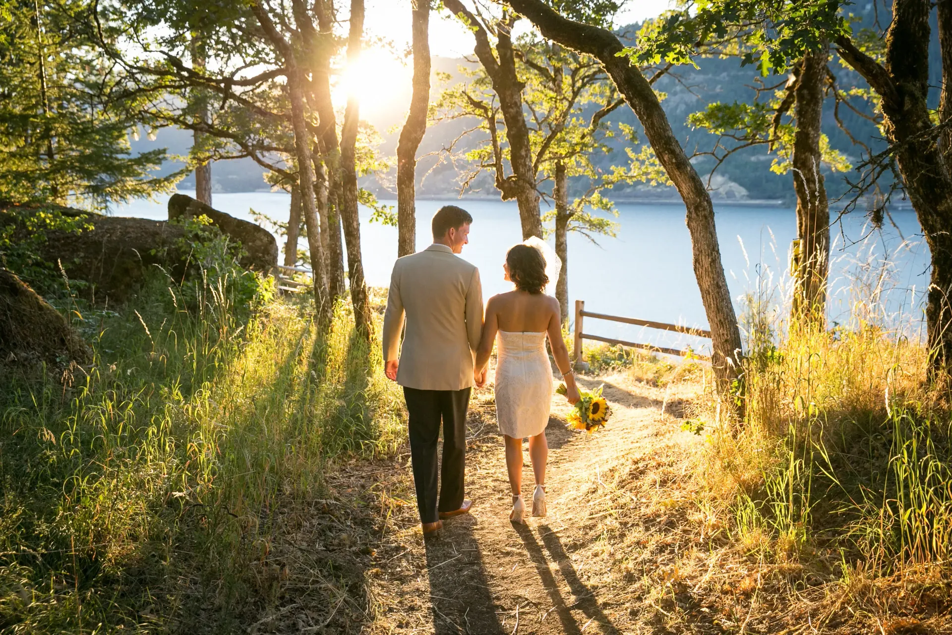 Couple holding hands walking on a path towards a lake, sun shining through trees.