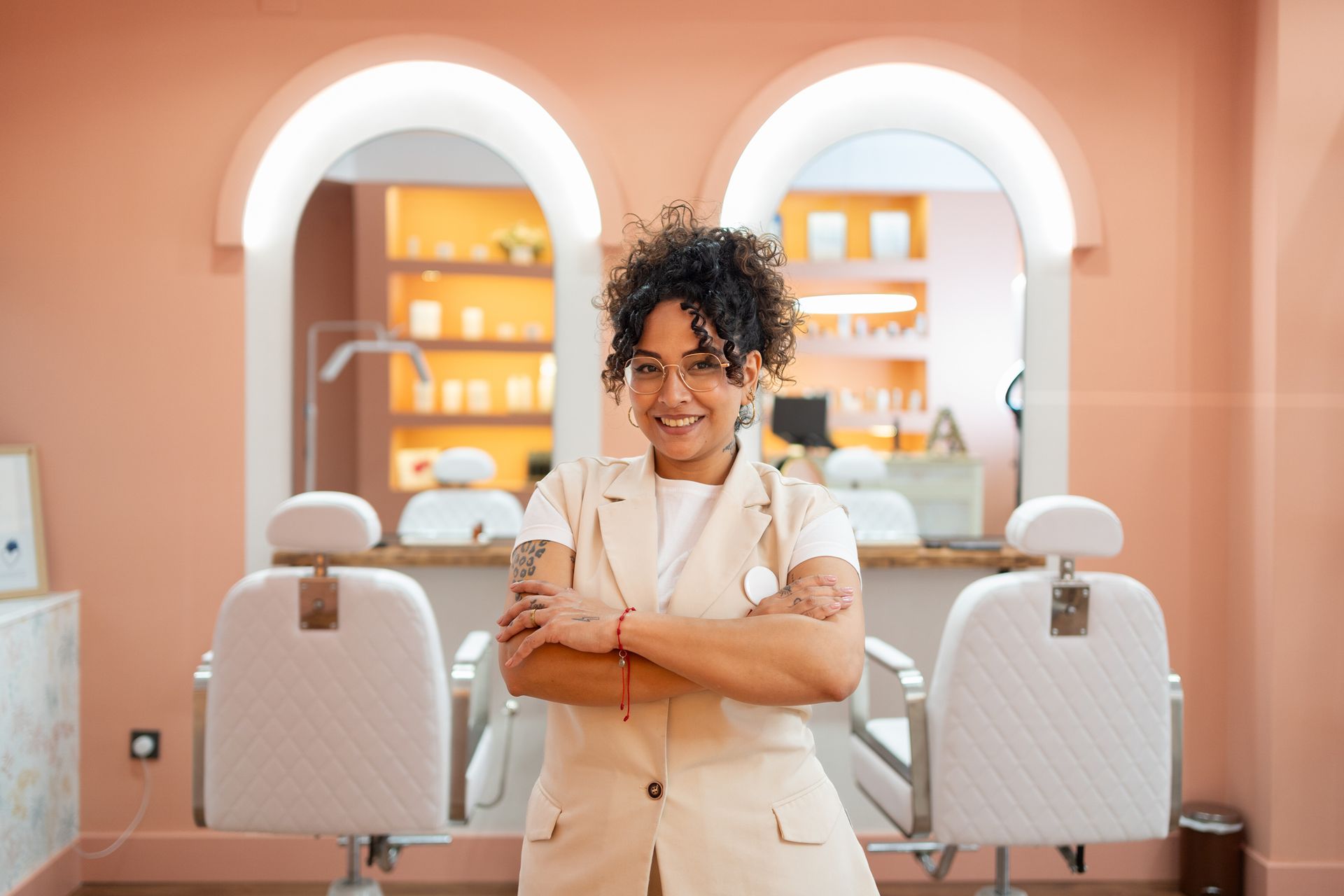 Woman with crossed arms smiles in a pink salon; barber chairs, mirrors, and products are visible.