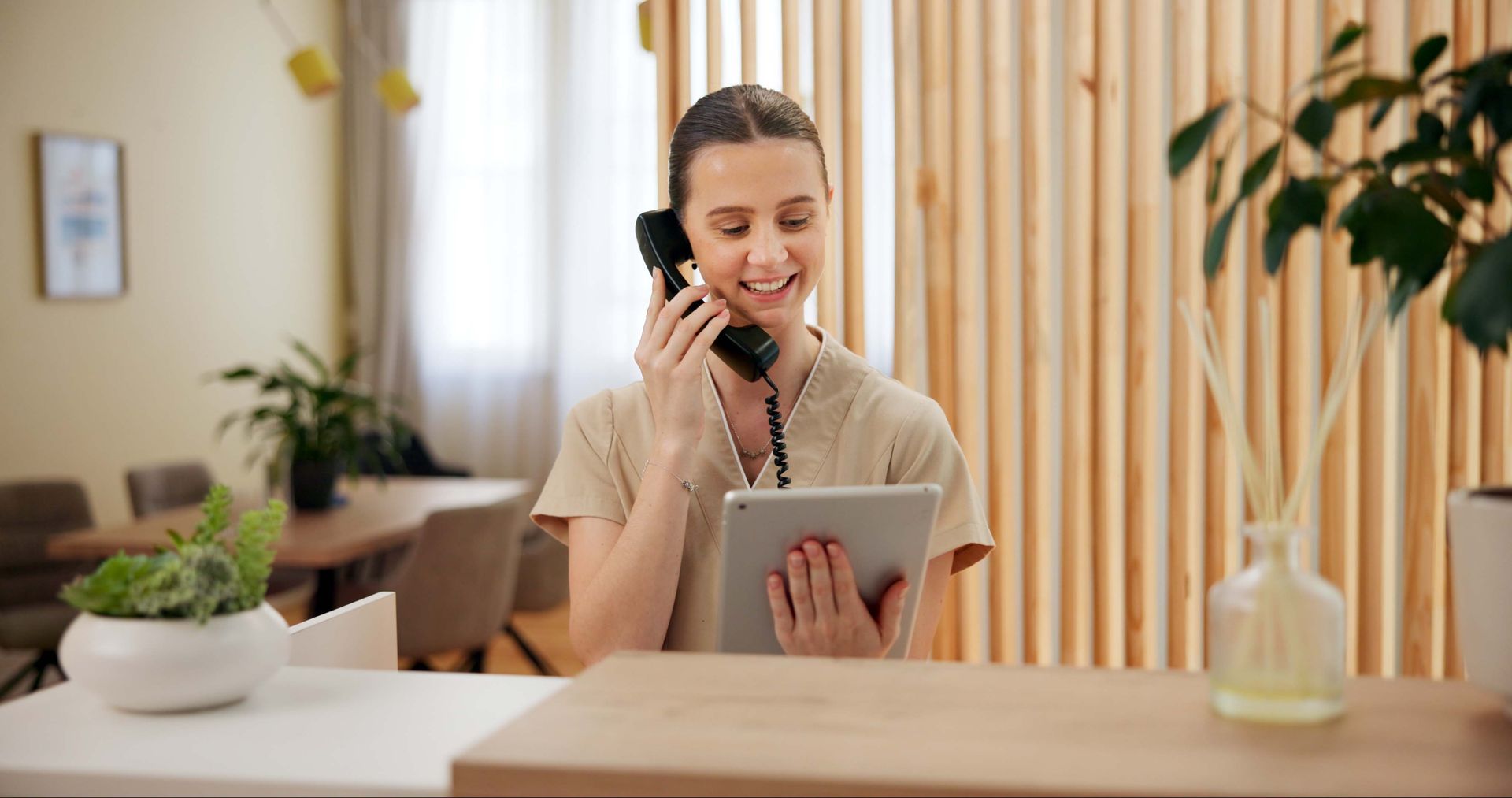 Woman smiling, talking on phone, holding tablet at a reception desk in a bright room.