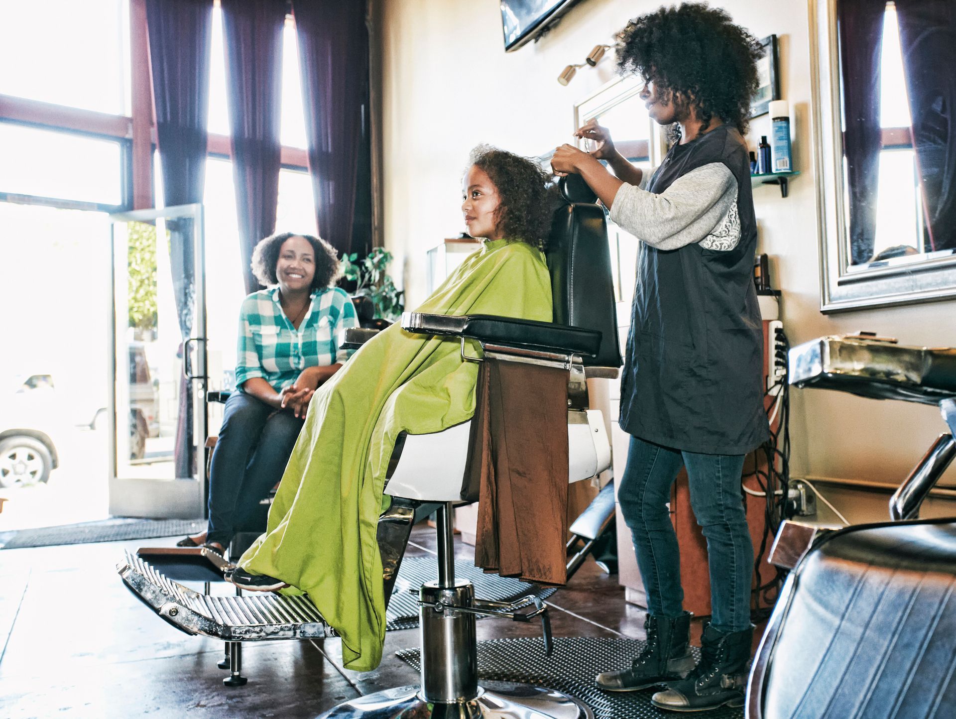 Woman styling a girl's hair in a salon. Another woman watches from a chair near an open door. Woman styling a girl's hair in a salon. Another woman watches from a chair near an open door.