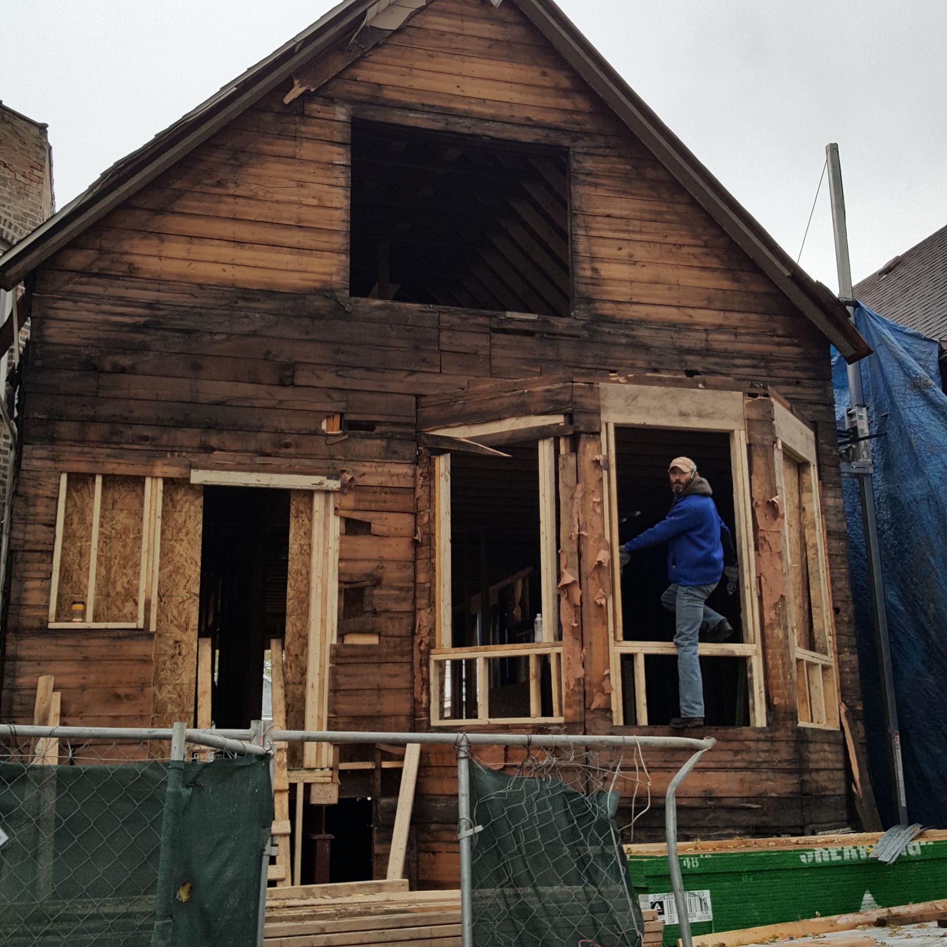 A man in a blue jacket stands in front of a wooden house under construction
