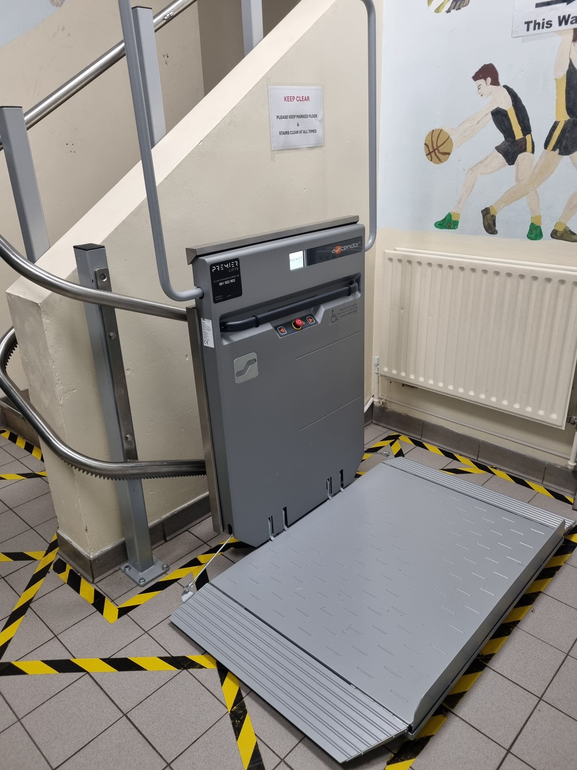 A grey inclined platform lift installed next to a staircase with yellow and black caution tape on the floor.