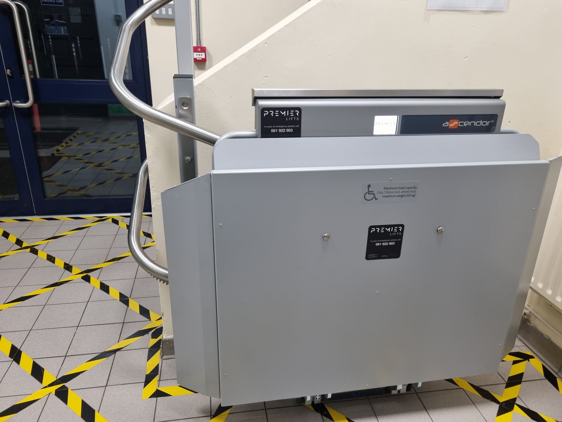 A gray wheelchair platform lift folded against a wall, situated near a doorway in a building with yellow safety tape.