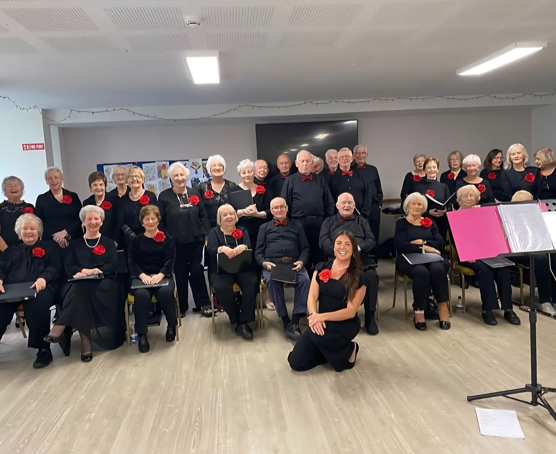 A group of people wearing black, many with red flowers pinned to their clothes, pose together in a bright, indoor room.