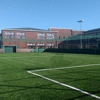 A bright green artificial turf soccer field with white boundary lines, set in front of a modern red brick school building.