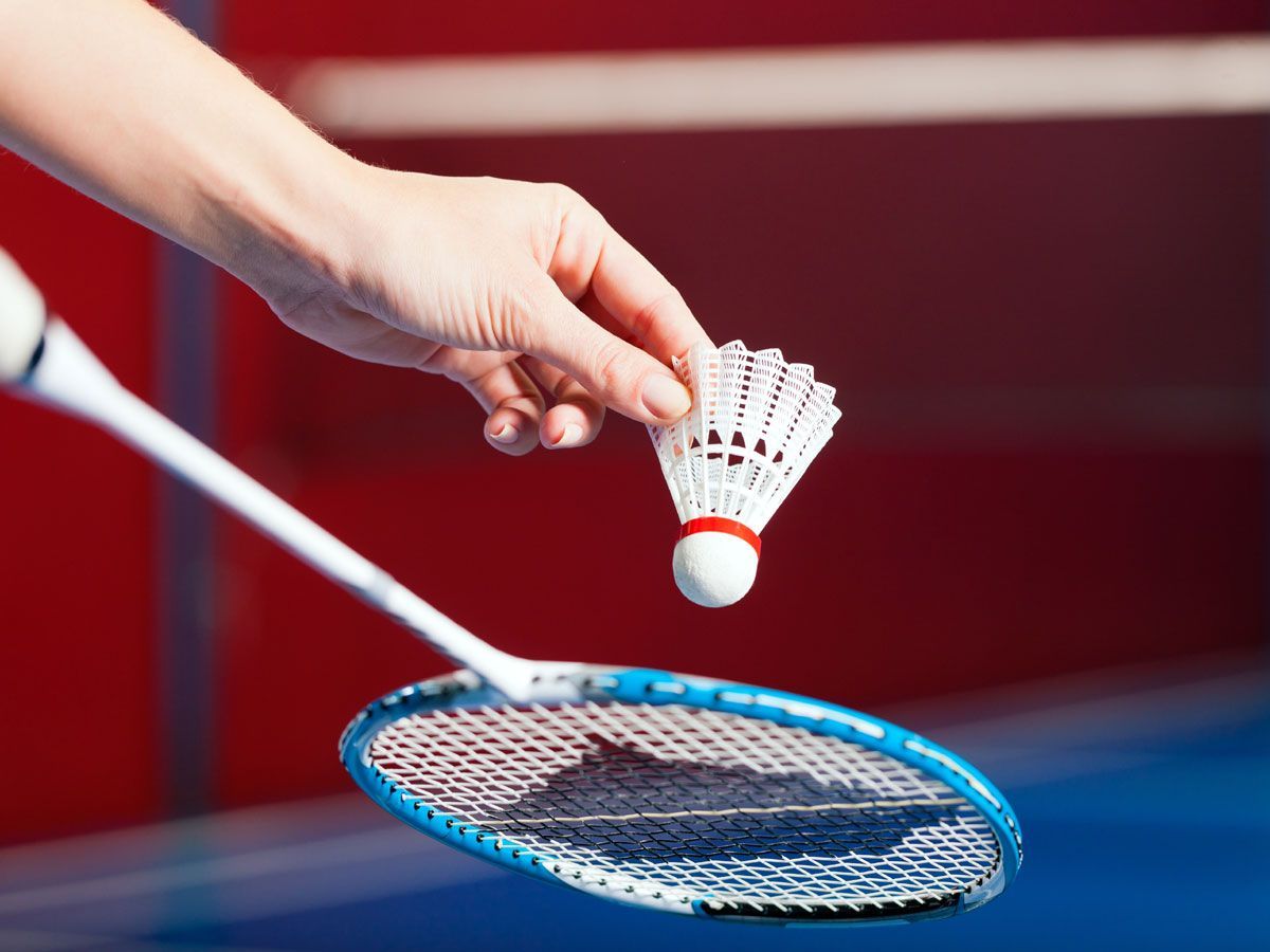 A hand holds a white badminton shuttlecock above a blue-framed racket, with a blurred sports background.