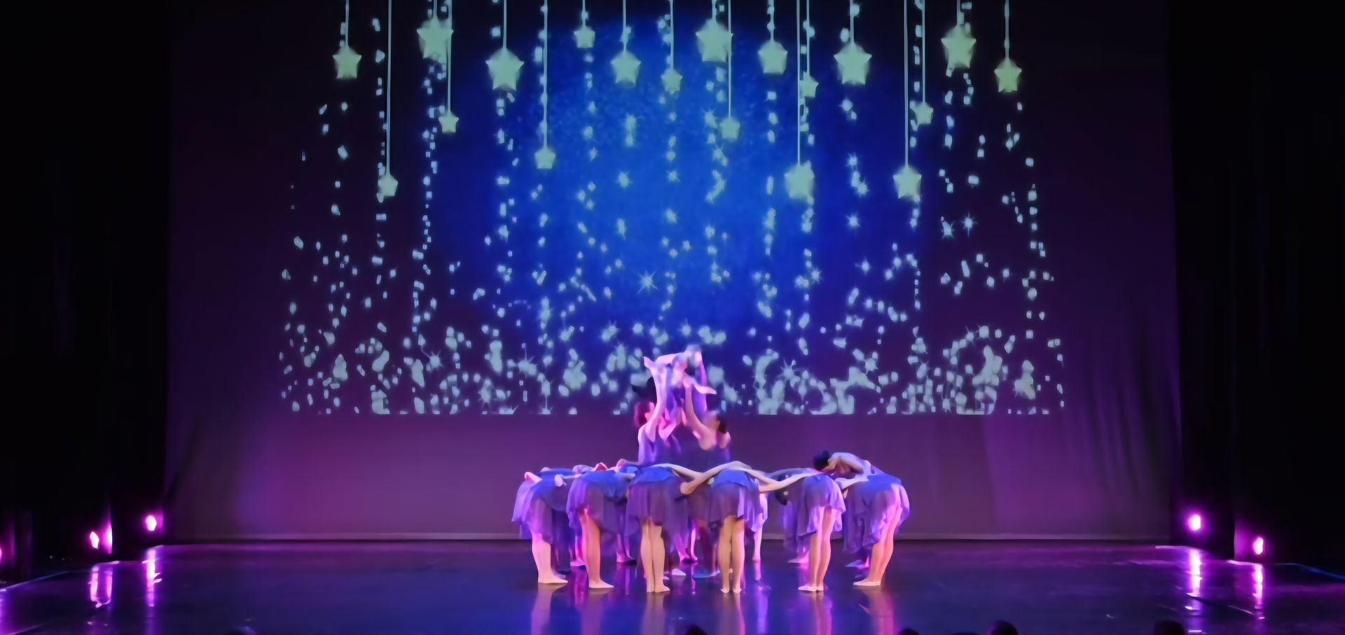 A group of dancers in purple costumes crouch together on a stage against a backdrop of glowing, hanging star lights.