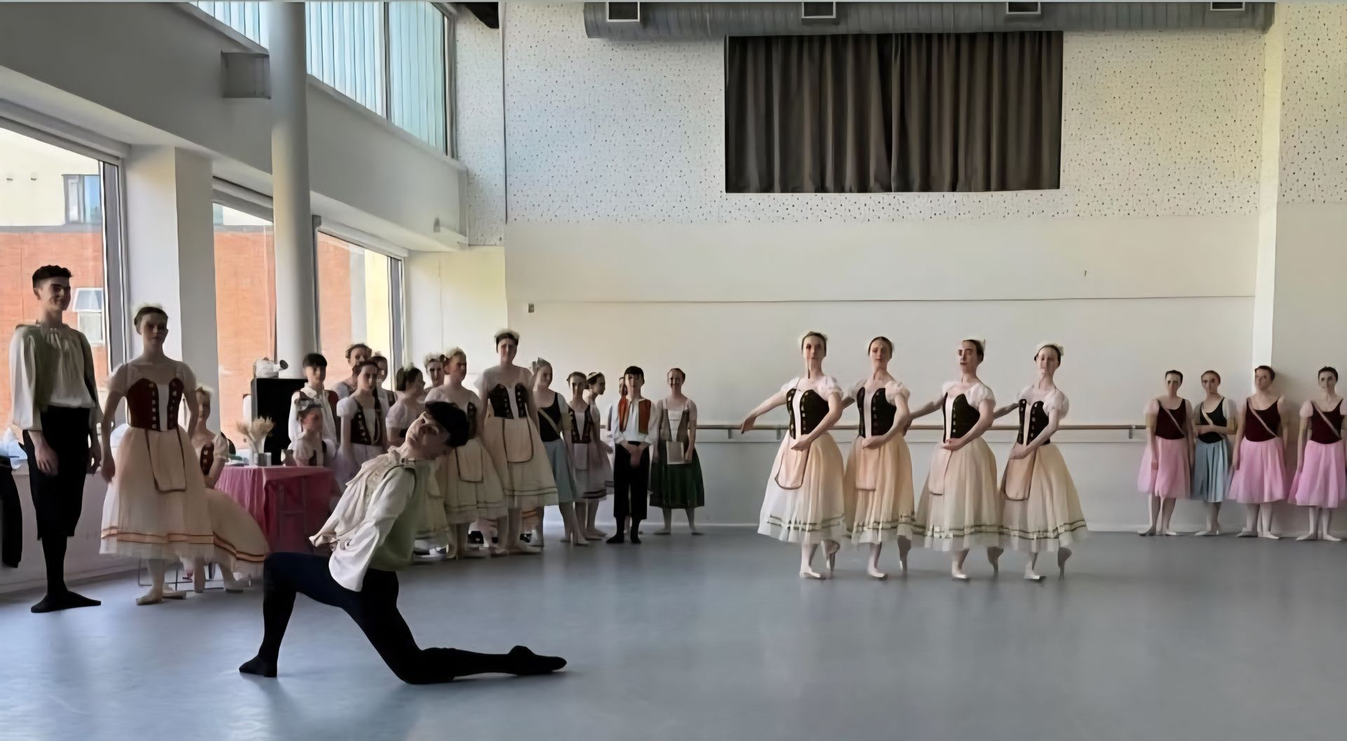 Ballet dancers perform a routine in a studio, with one dancer kneeling in the foreground and a line of dancers behind.