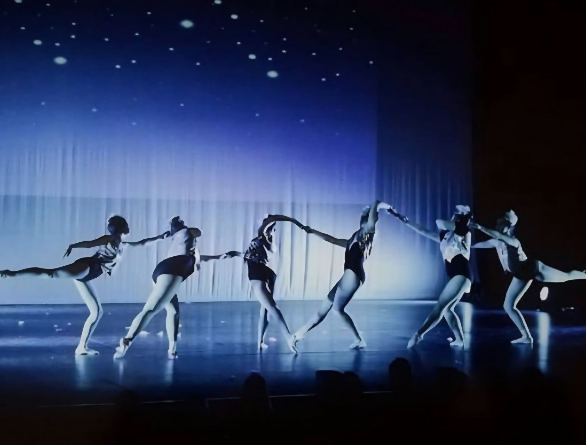 Six dancers in dark leotards perform a choreographed routine in a line against a backdrop with a starry night projection.