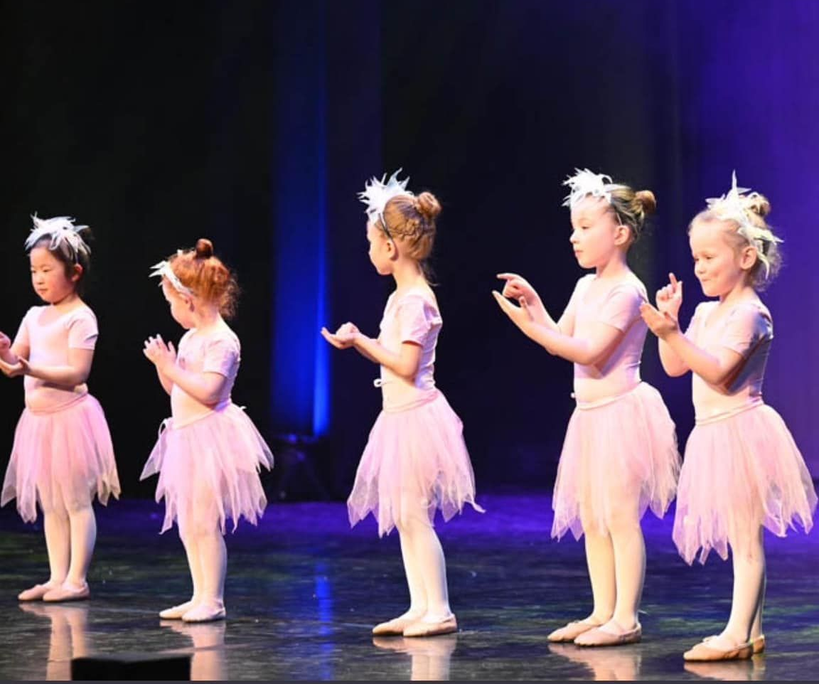 Five young dancers in pink tutus and hair accessories perform on a stage with a dark background.