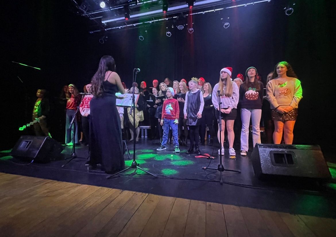 A choir performs on a stage under colorful stage lights, led by a conductor seen from behind.