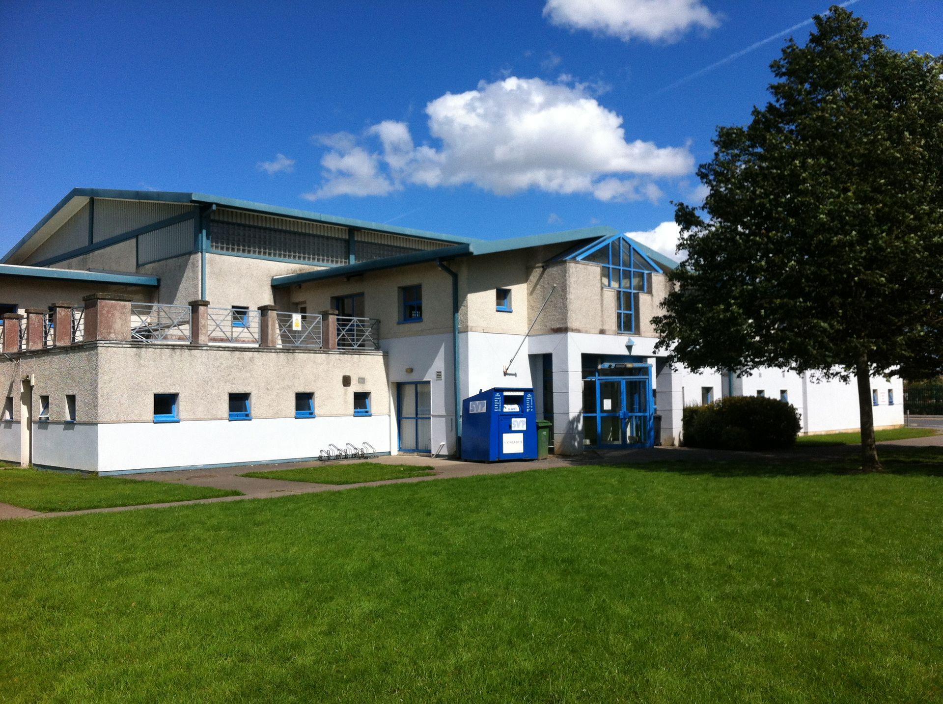 A two-story building with white walls, a blue trim, and a large metal roof, situated next to a tree and a grassy lawn.