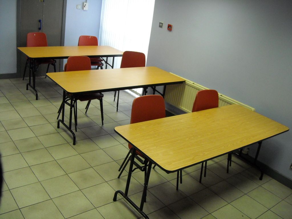 A room with three light-wood rectangular tables arranged in a row, each featuring two orange chairs on a tiled floor.