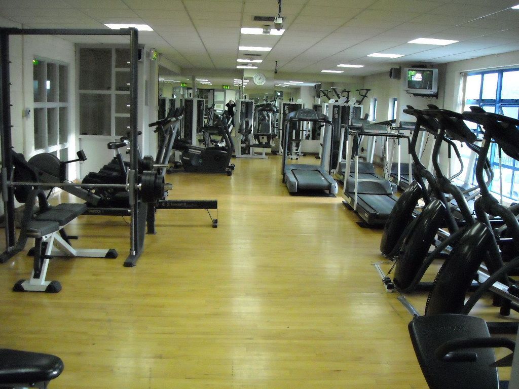 An empty gym with rows of cardio equipment, weight benches, and treadmills on a wooden floor, viewed from the hallway.