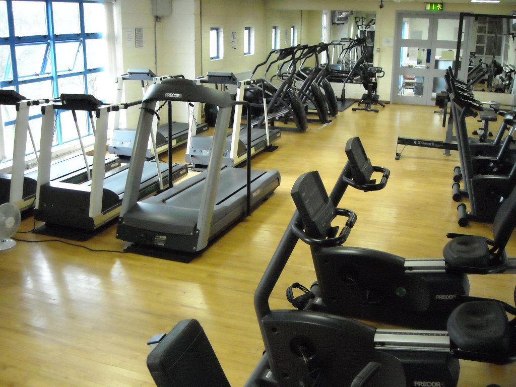 A gym with multiple treadmills, elliptical machines, and stationary bikes arranged on a light-colored wood floor.