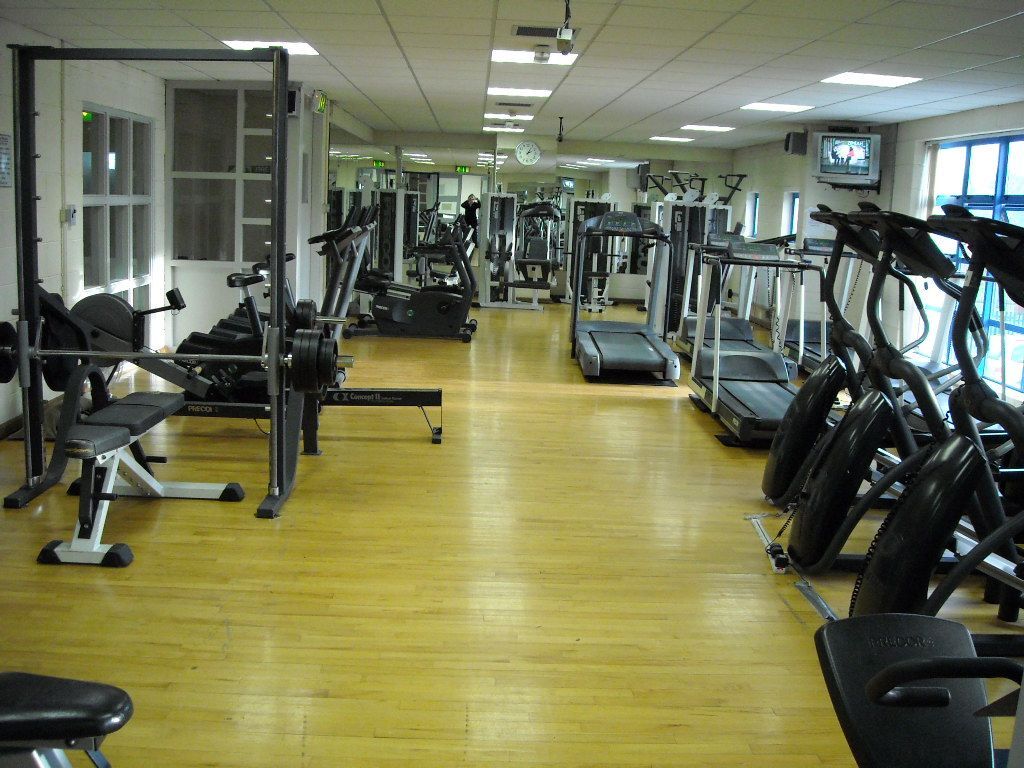 An interior view of a gym featuring rows of treadmills, exercise bikes, and weightlifting equipment on a wooden floor.