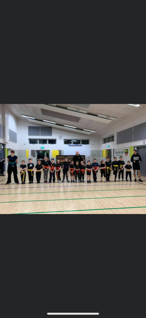 A group of children in martial arts uniforms stand in a line on a wooden gym floor, with instructors at the ends.