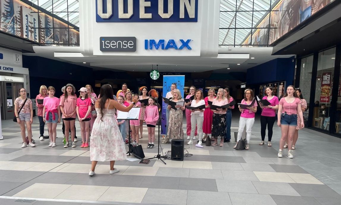 A group of singers performing in front of an Odeon cinema, led by a conductor standing with sheet music on a stand.