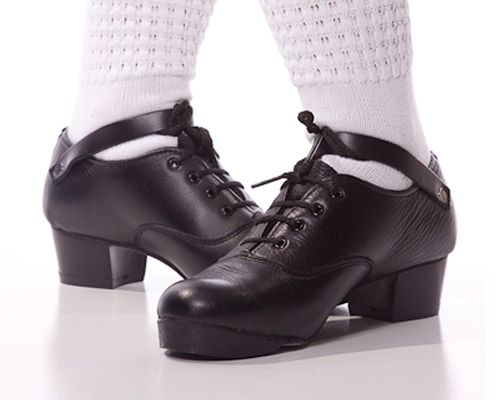 A pair of black leather character tap shoes with lace-up fronts and ankle straps, worn with white leg warmers.