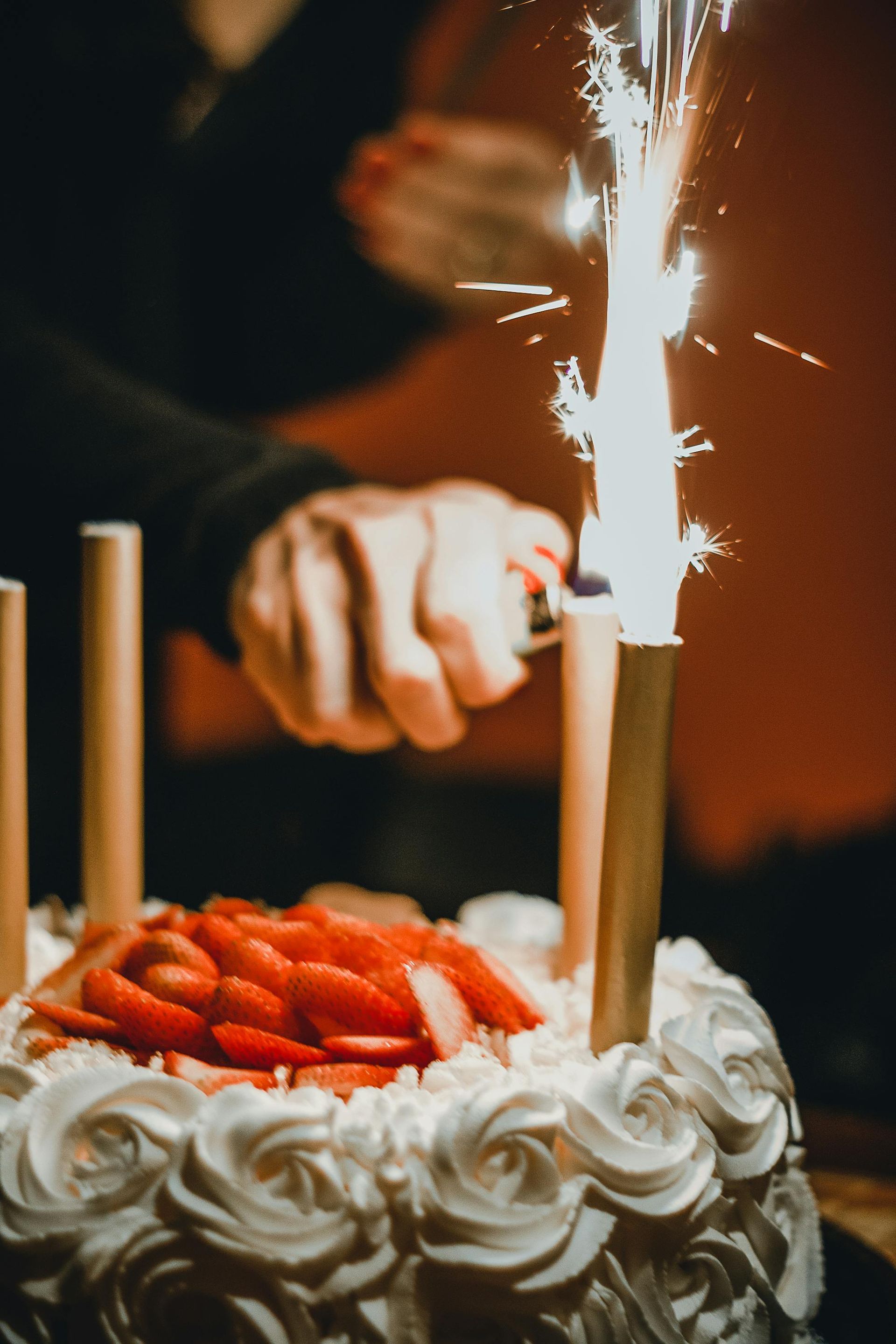 A hand uses a lighter to ignite a sparkler on a strawberry-topped white frosted cake.
