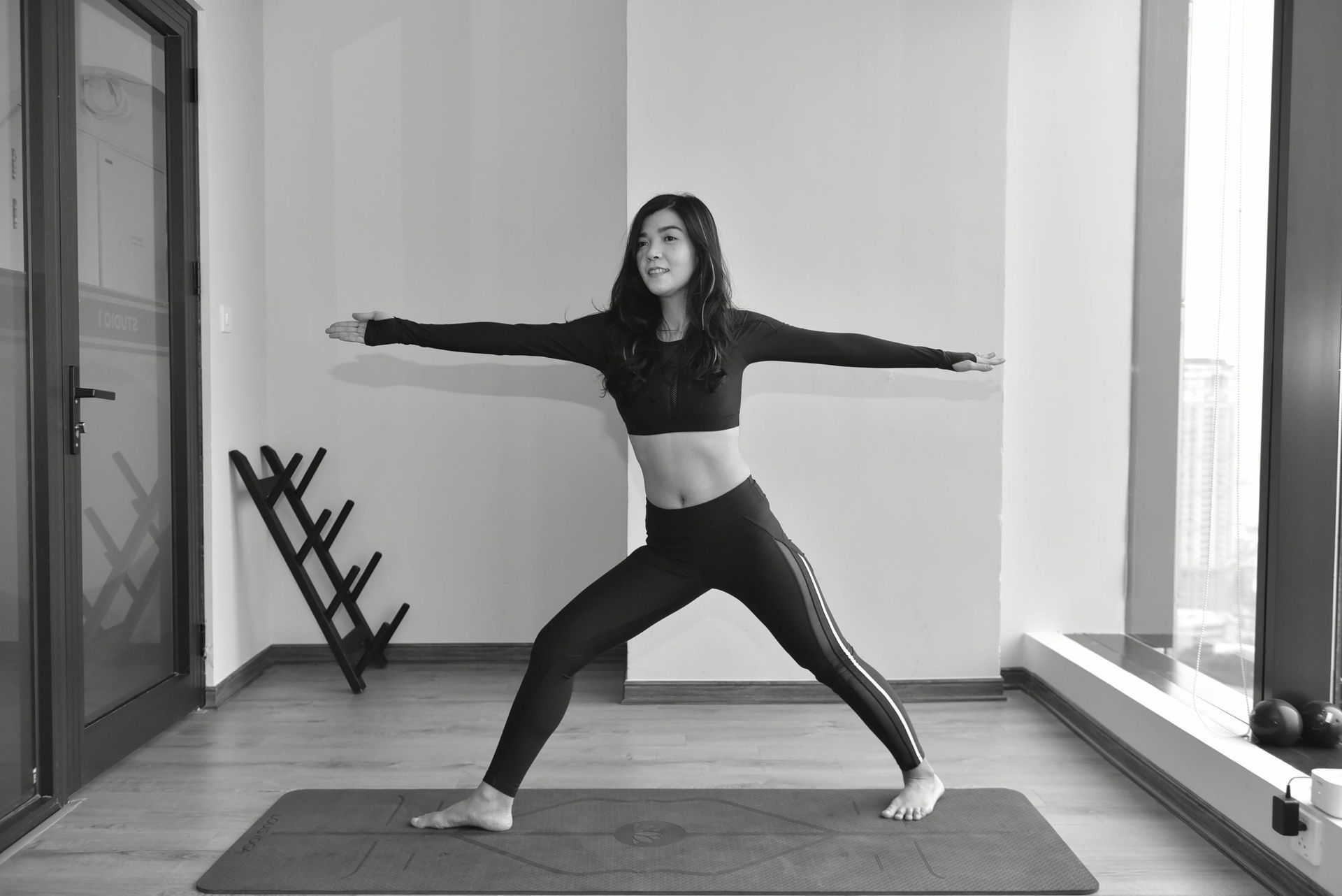 A person in black athletic wear practices the warrior II yoga pose on a mat in a bright, modern room.