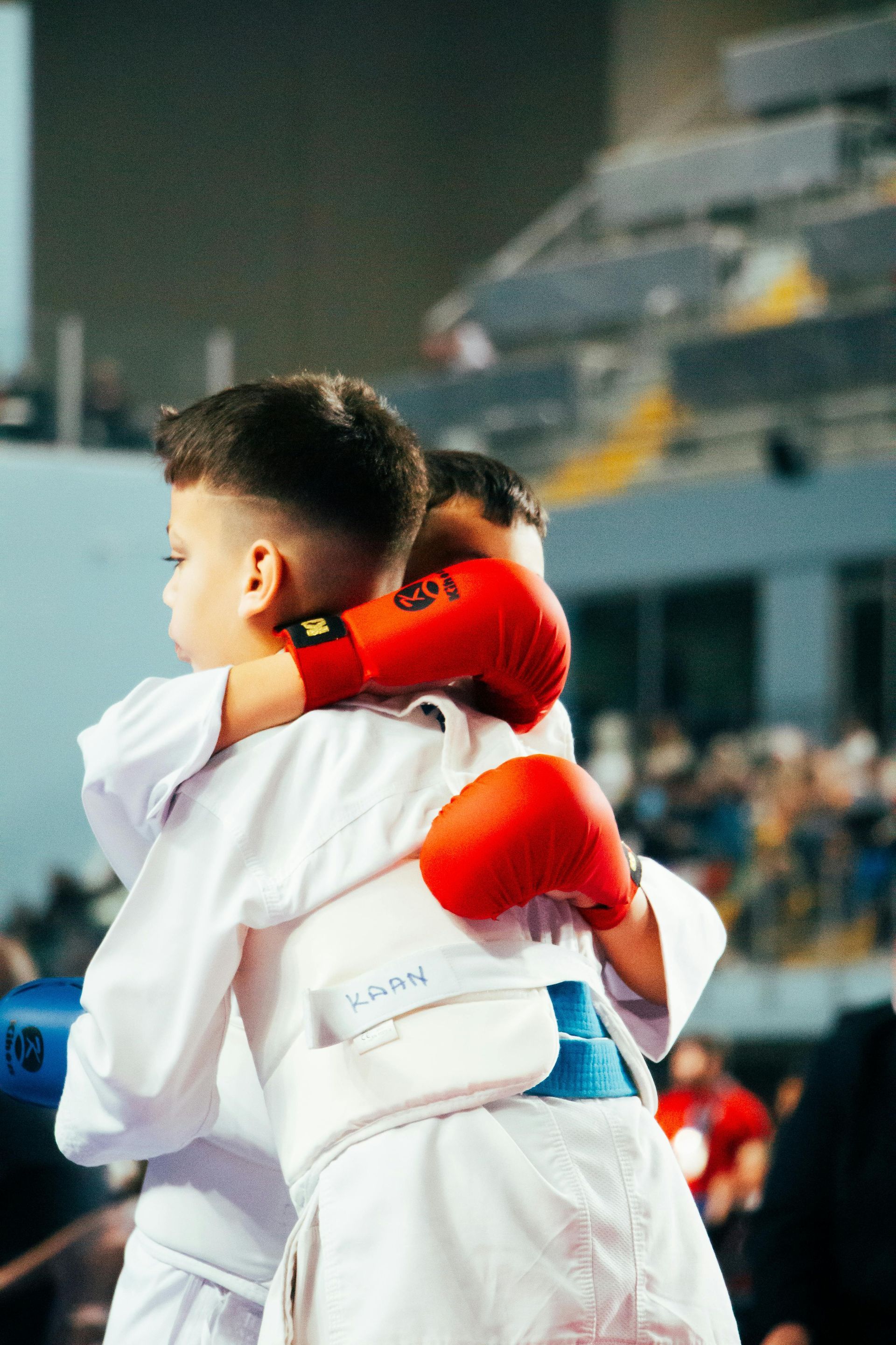 Two karate competitors in white uniforms and red sparring gloves embrace after a match in an indoor arena.