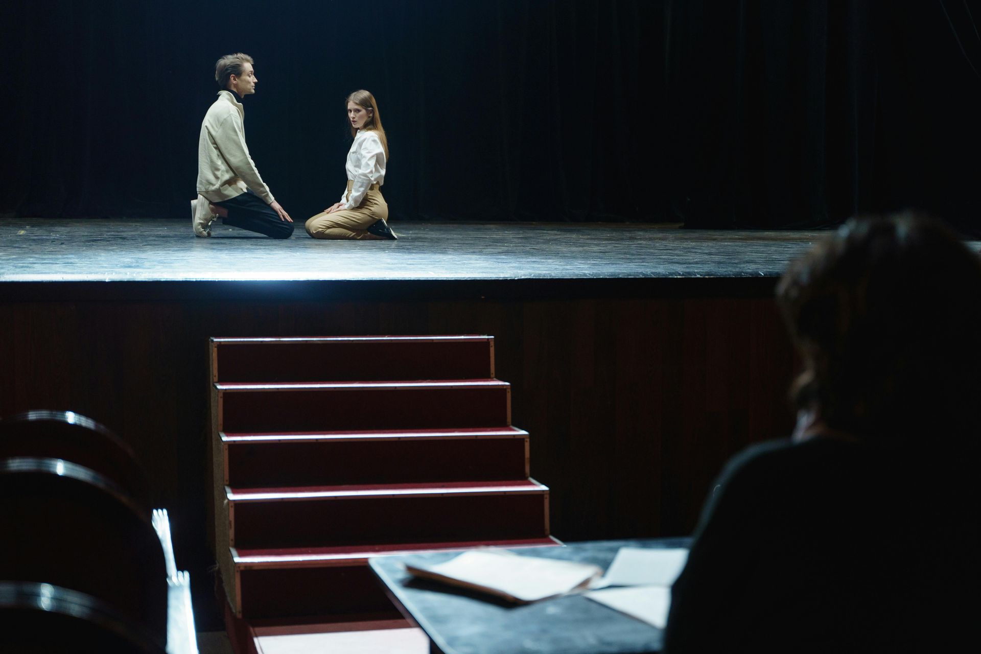 Two people kneel on a theater stage, viewed from the audience with a table and stairs in the foreground.
