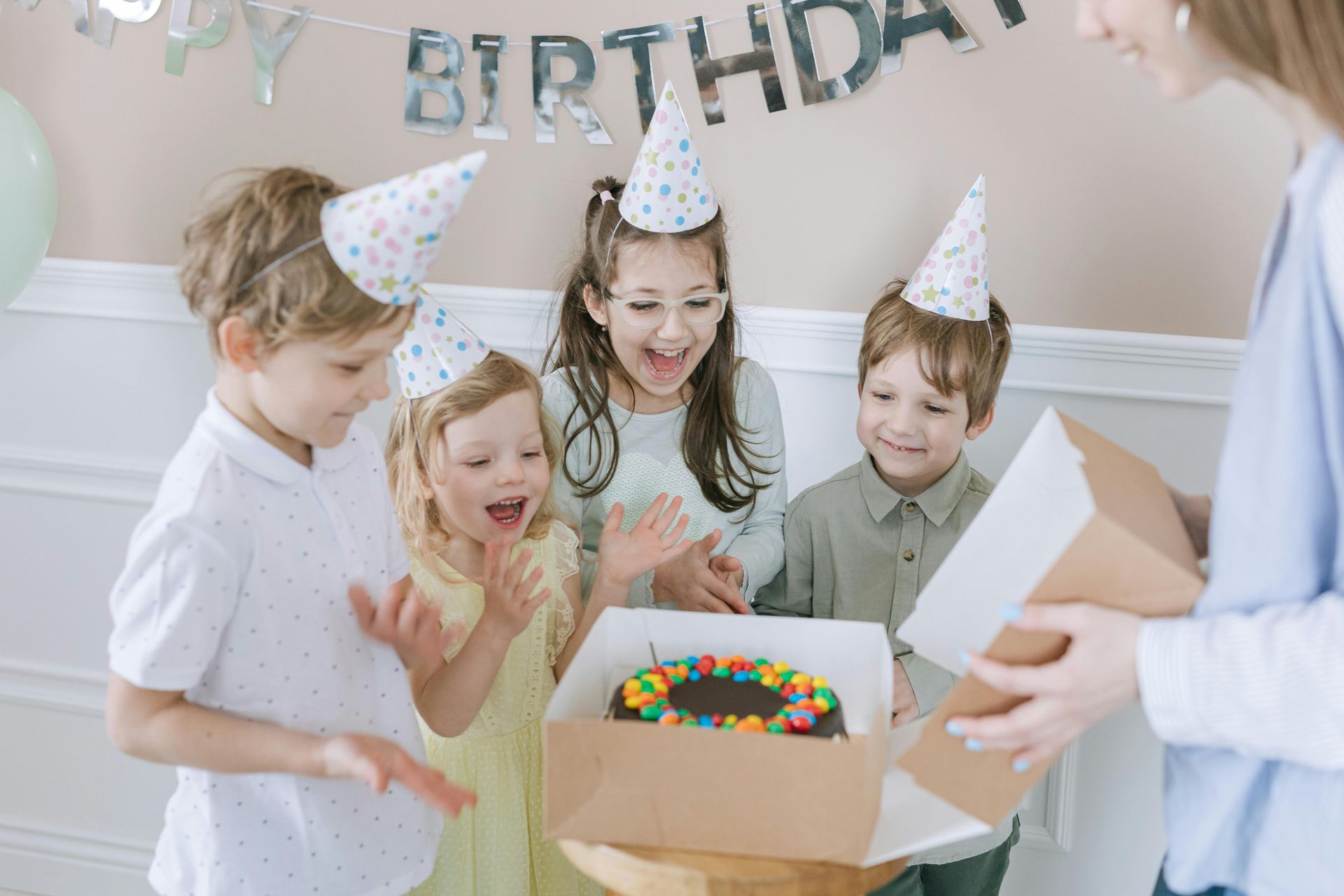 Four children wearing party hats gather around a birthday cake in a box while an adult opens it.