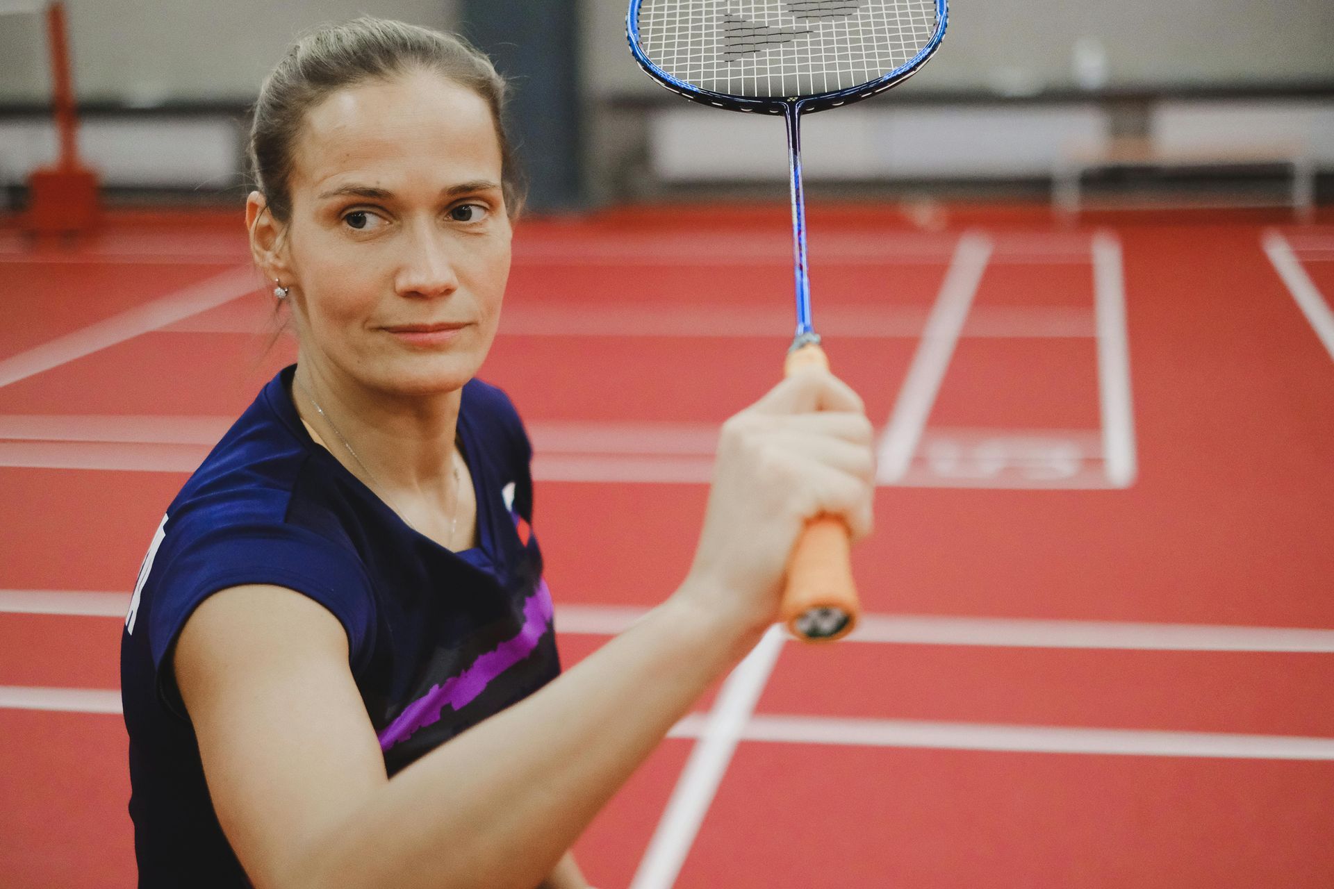 A badminton player holds a racket on a red court, looking forward with a focused expression.