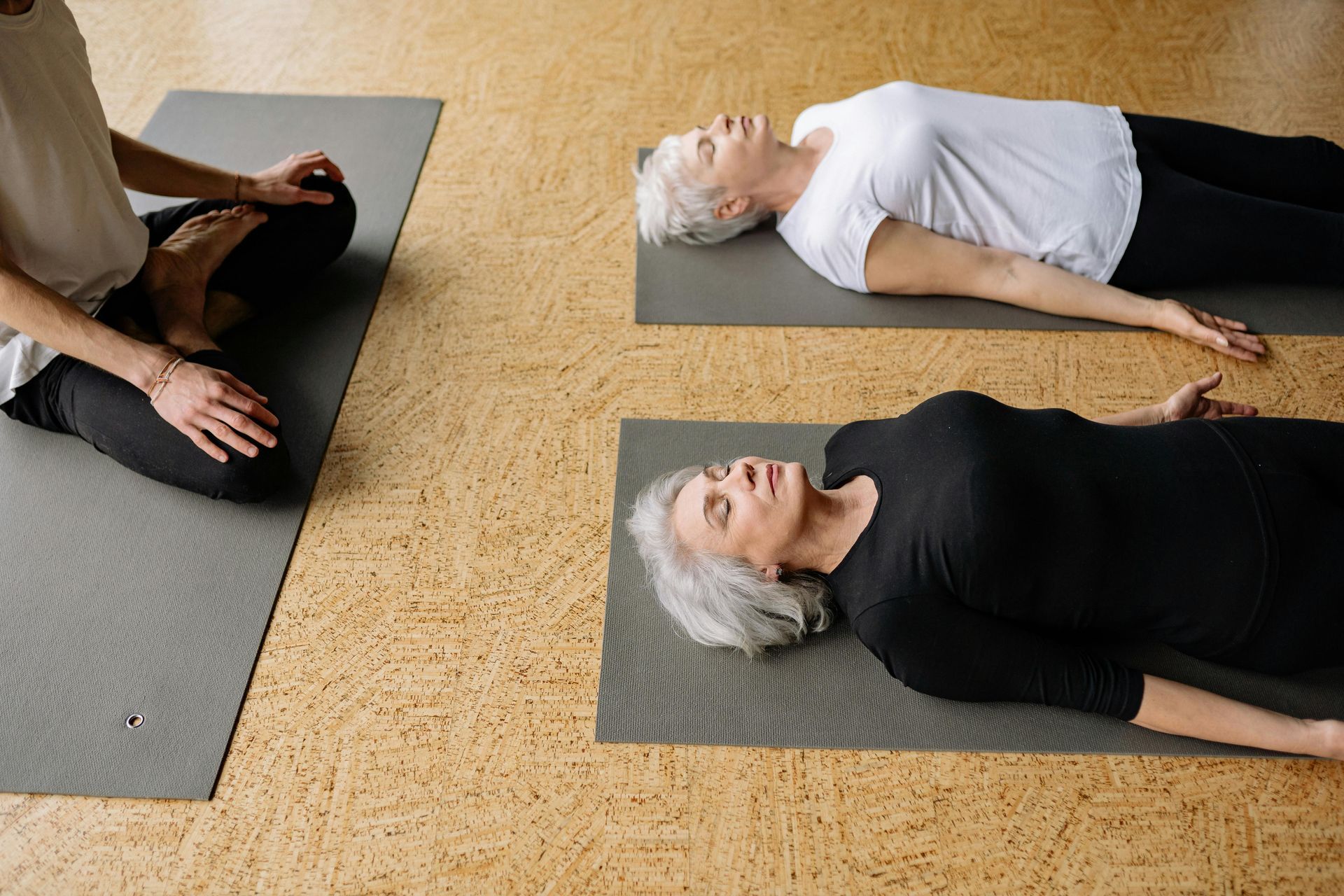 A person leads a yoga class, sitting cross-legged as two people lie flat on mats on a wooden floor in Savasana pose.