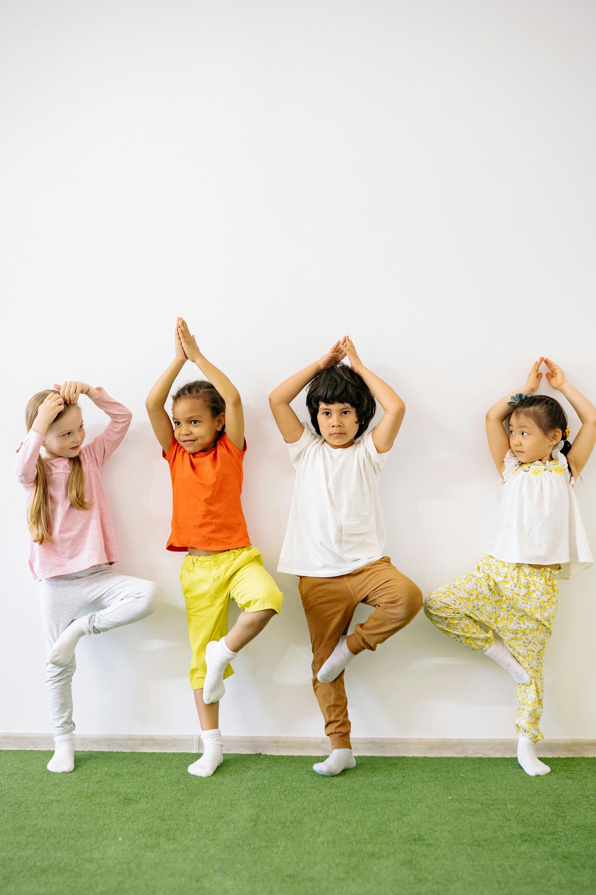 Four children stand against a white wall on green turf, balancing on one leg with hands joined above their heads.