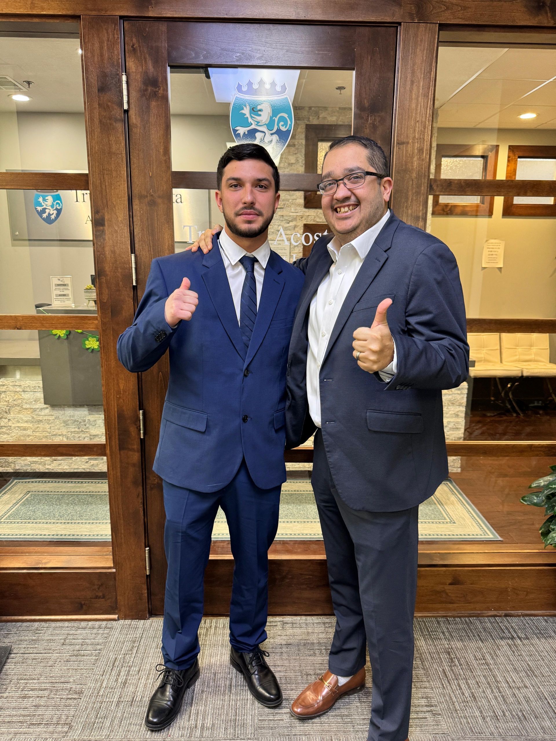 Two men in suits give thumbs up, inside a building with a wooden door and a patterned carpet.