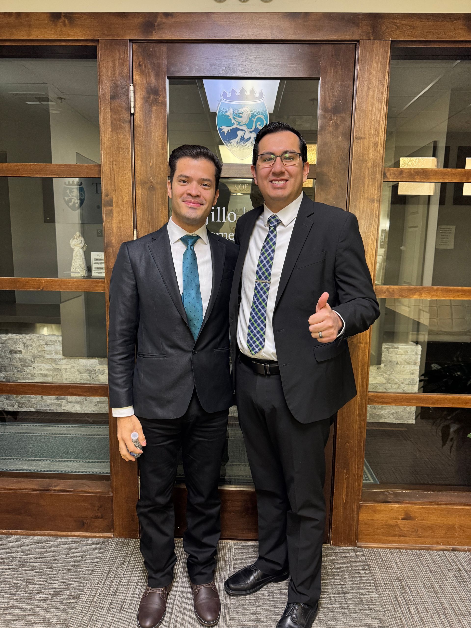 Two men in suits stand in front of a wood door. One gives a thumbs up; both smile.