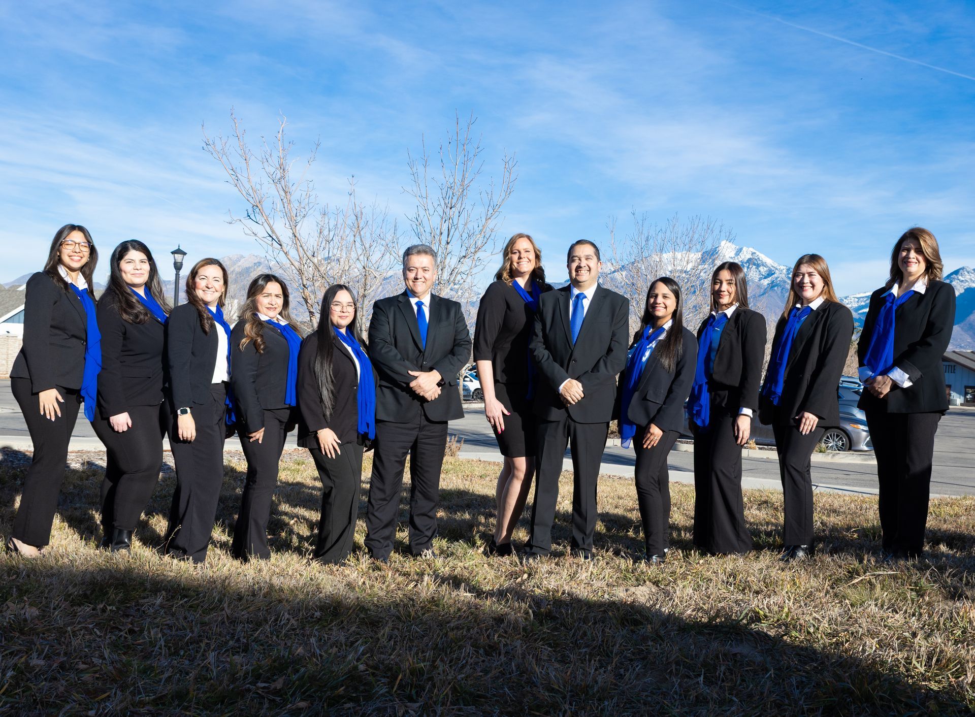 Group of people in business attire, outdoors, posing in front of mountains and a blue sky, wearing blue scarves.