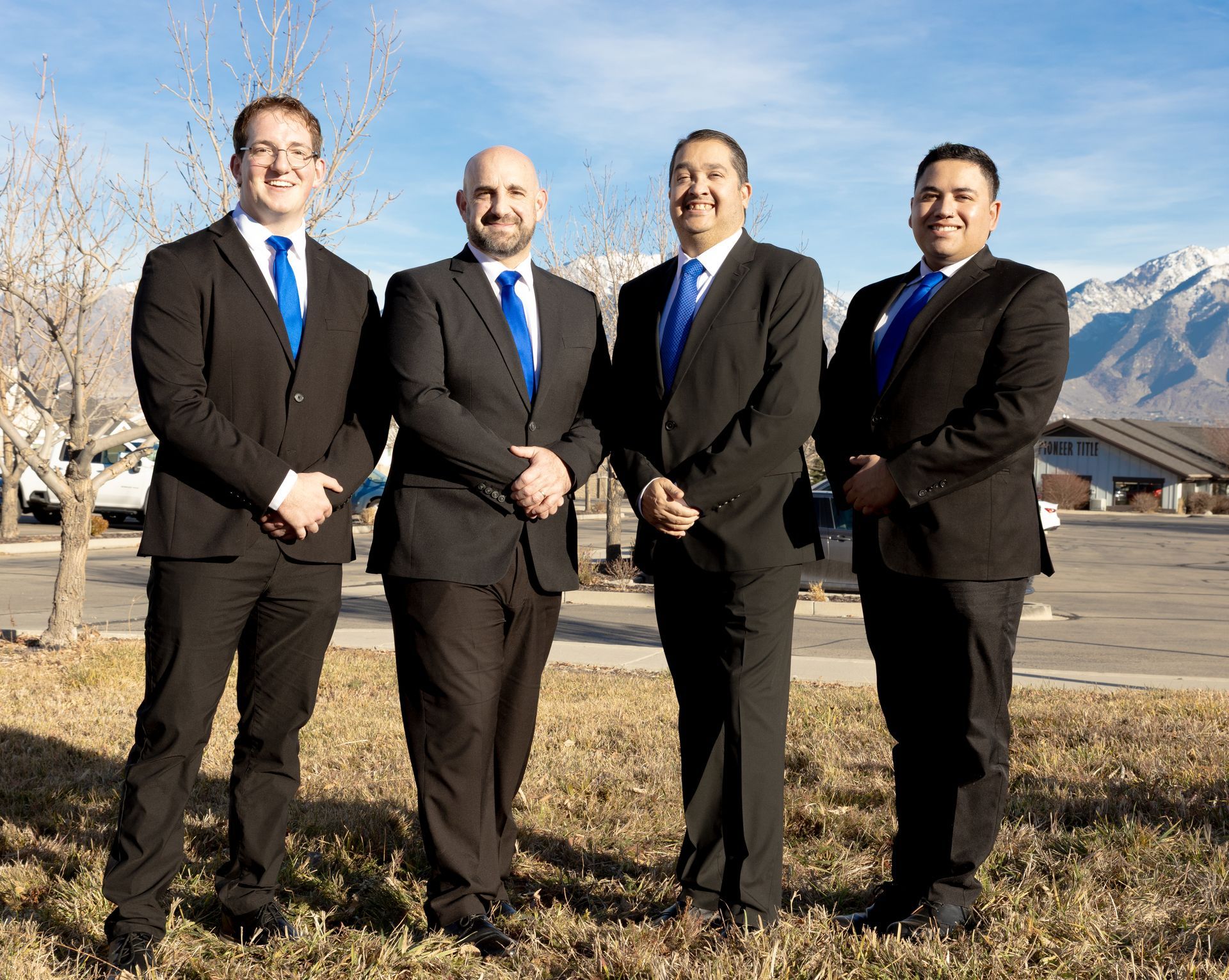 Four men in suits and blue ties pose outside with mountains in the background.