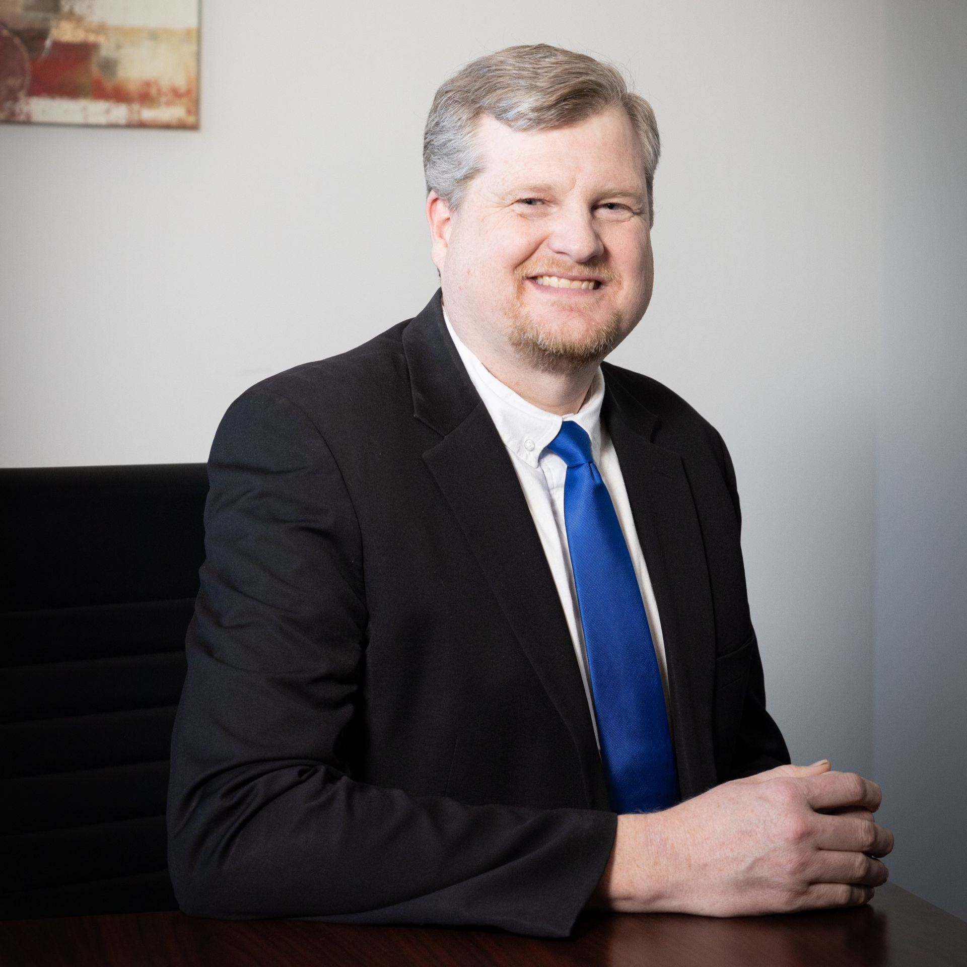 Man in black suit and blue tie sitting at a desk, looking at the viewer.