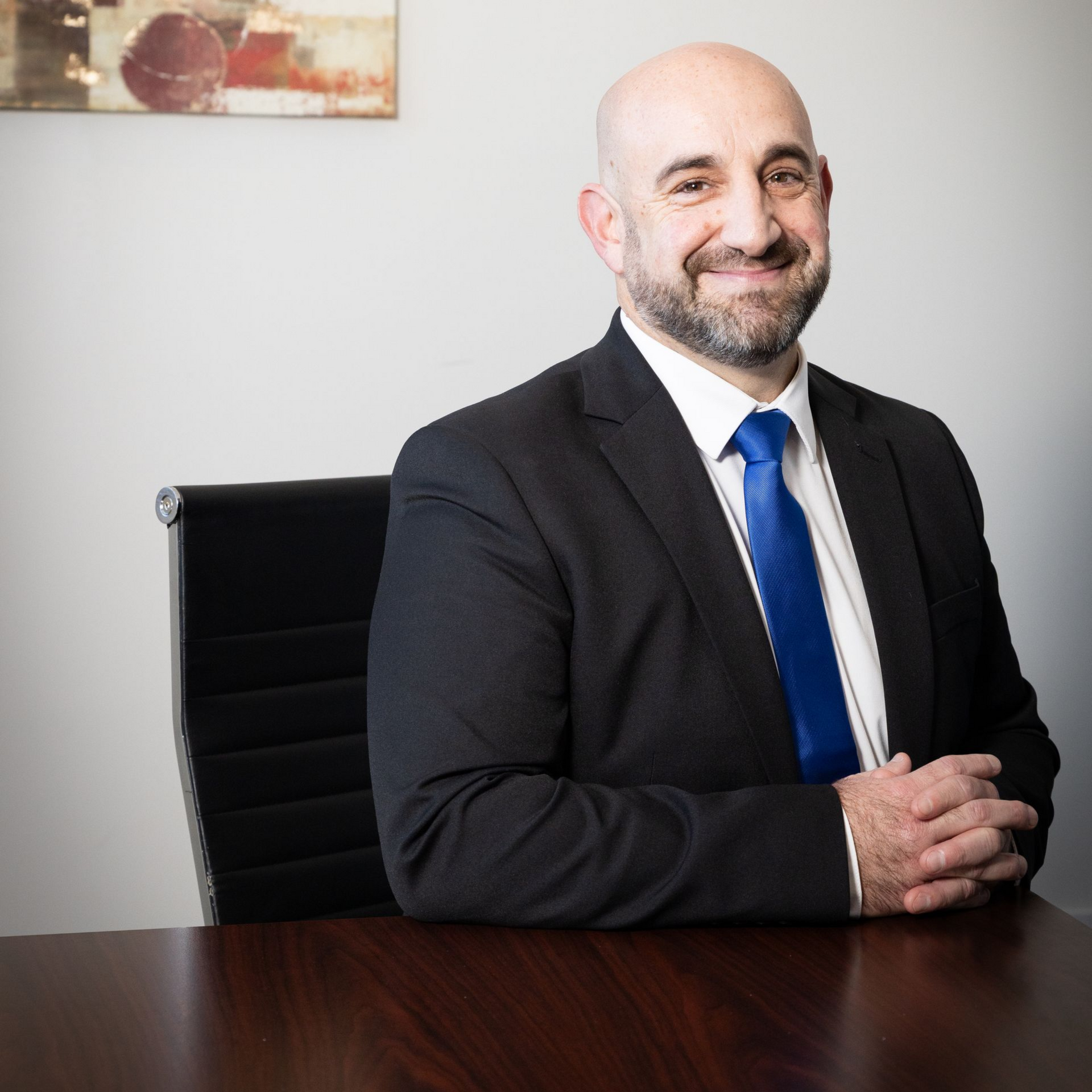 Bald man in suit, blue tie, smiling, seated at a desk.