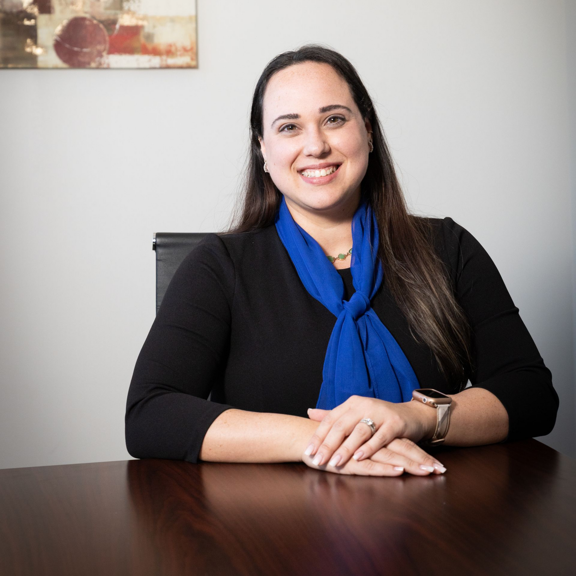 Woman seated at a desk, smiling with folded hands, wearing a black shirt and blue scarf.