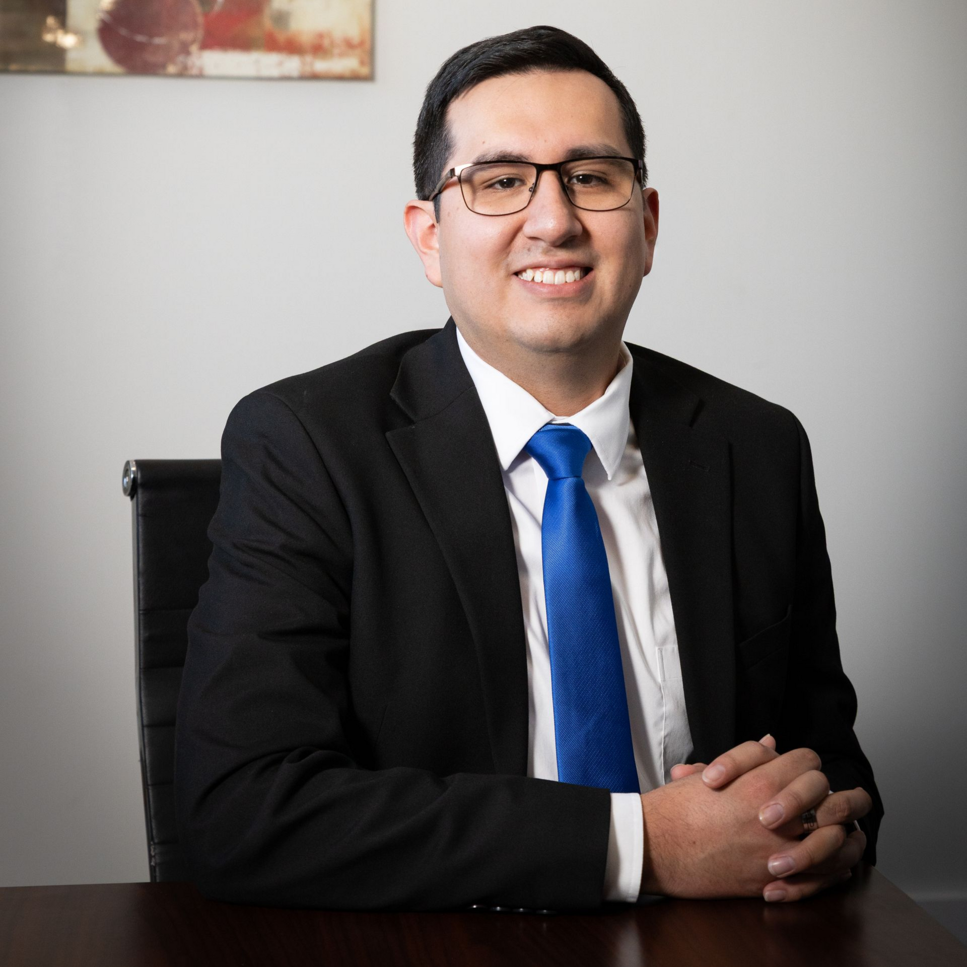 Man in suit, smiling, seated at a desk, looking at the viewer. White shirt, blue tie.