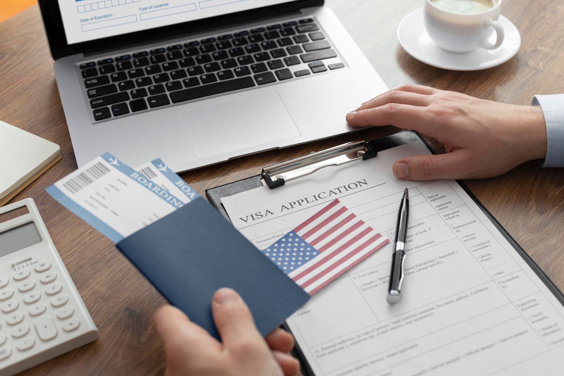 Hands holding a passport with boarding passes, US visa application, laptop, and calculator on desk.