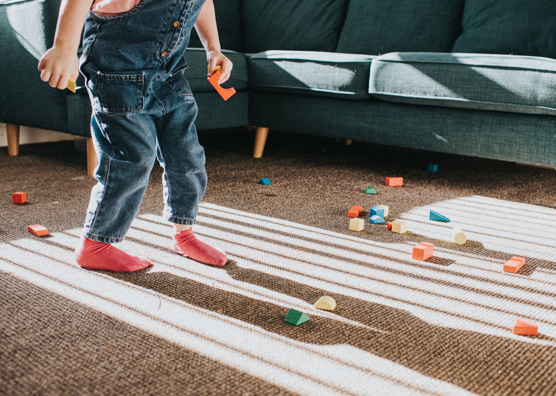 Child in denim overalls and pink socks plays with blocks scattered on carpet near a sofa.
